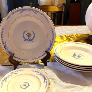 May include: A set of white porcelain plates with a blue and gold leaf design. The plates have a gold rim and a central floral emblem. Several plates are stacked, and others are displayed on wooden stands.