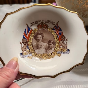 May include: A white, scalloped-edge dish with a gold rim. The center features a portrait of Queen Elizabeth II, crowned June 2nd, 1953, flanked by flags and lions. The dish has the words "Dieu et mon droit".