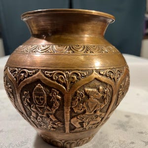 Pode incluir: Um vaso decorativo de cor cobre, com uma abertura larga e uma forma arredondada. O vaso apresenta entalhes em relevo detalhados de figuras e motivos florais. O design sugere um artefato tradicional ou cultural.