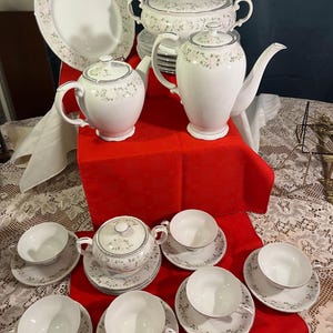 May include: A white porcelain tea set with a floral pattern and silver trim. The set includes a teapot, coffee pot, sugar bowl, serving bowl, and several teacups and saucers. The set is displayed on a red tablecloth.