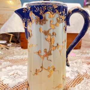 以下が含まれることがあります： 背の高い円筒形のアンティーク磁器コーヒーポット。クリーム色のボディに金色の透かし模様、ハンドル、蓋、ベースに濃い青色のアクセントが施されています。注ぎ口と小さなノブが付いた蓋があります。