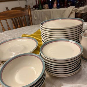 May include: A collection of off-white ceramic dinnerware with a teal and mauve border. The set includes plates and bowls, stacked in neat piles on a table. The tableware is arranged on a white tablecloth, suggesting a dining setting.