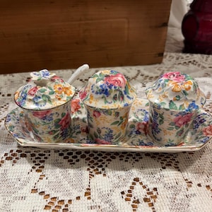 May include: A floral-patterned ceramic condiment set featuring three small lidded containers and a spoon, all resting on a matching rectangular tray. The design includes pink, blue, and yellow flowers on a white background, with gold trim.