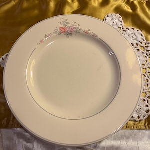May include: A set of off-white ceramic plates with a delicate floral design. The plates have a thin gray border and a decorative arrangement of pink and red roses with green leaves. The plates are stacked on a gold and white surface.