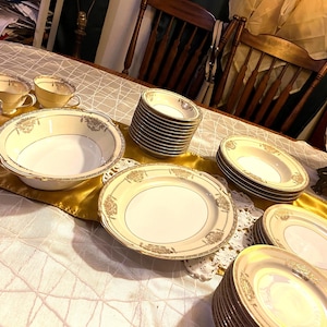 Puede incluir: Una colección de porcelana crema vintage con ribetes dorados y detalles florales. El juego incluye tazas de té, cuencos y platos de varios tamaños, dispuestos sobre una mesa con un camino de mesa dorado y un mantel blanco.