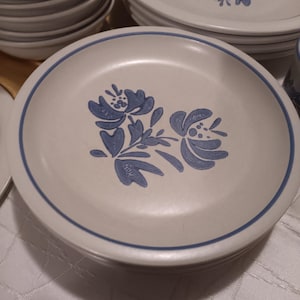 Puede incluir: Una pila de platos de cerámica blanquecina con un diseño floral azul y un borde azul. Se ven otros platos y cuencos en el fondo, lo que sugiere un juego de vajilla completo. Los platos tienen una estética clásica y vintage.
