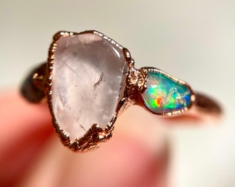 May include: A rose gold ring featuring a clear quartz and an iridescent opal gemstone. The ring is crafted in a wire wrapped style.