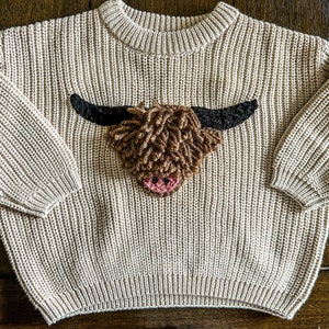 Op de afbeelding: Een beige gebreide trui met een bruine en zwarte applicatie van een Schotse hooglander.