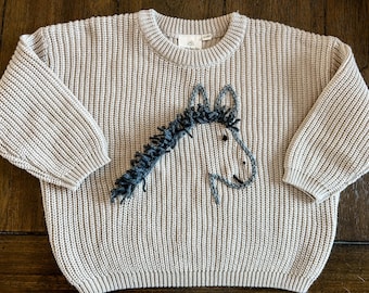 May include: A beige knitted sweater with a gray embroidered horse design on the front.
