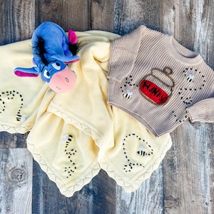 Op de afbeelding: Een beige baby trui met een honingpot en bijen ontwerp, een gele babydeken met bijen borduursel en een blauw pluche ezel speelgoed worden gepresenteerd op een houten oppervlak.