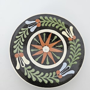 May include: A round ceramic plate with a black background featuring a central orange starburst design surrounded by a circular border. The outer rim is decorated with a repeating pattern of green leafy vines and light blue and cream-colored flowers.