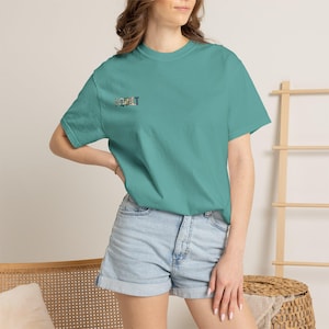 May include: A teal short-sleeve t-shirt featuring the text "I QUILT" printed on the left chest area. The letters are filled with a multi-colored, patterned design. The shirt has a crew neckline and a standard fit, paired with light blue denim shorts.