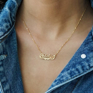 Puede incluir: Un collar de oro con un colgante personalizado en forma de placa con el nombre "Natalie" y dos pequeños corazones debajo del nombre. El collar lo lleva una persona que lleva una chaqueta vaquera.