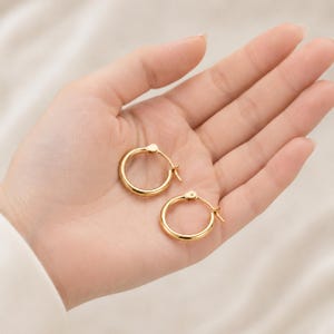 以下が含まれることがあります： 2つのゴールドカラーのフープピアス。ピアスは丸く、シンプルでクラシックなデザインです。ピアスは、柔らかくニュートラルな背景に対して、手で持たれています。