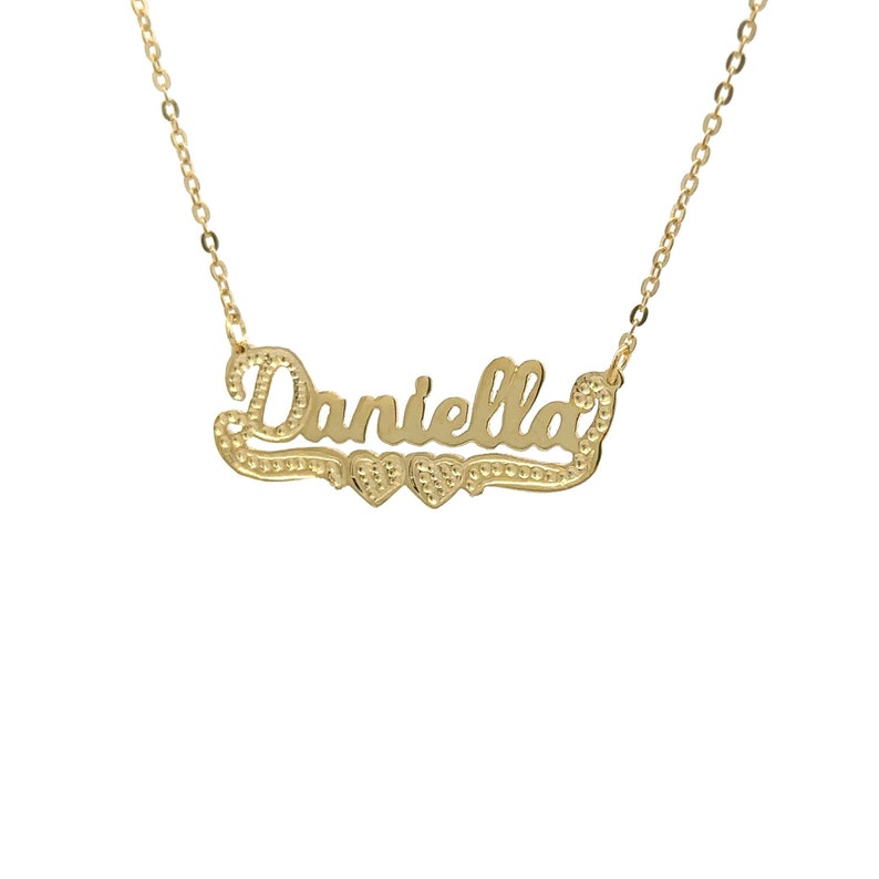 May include: Gold necklace featuring the name "Daniella" in a cursive script with two small hearts on either side of the name.