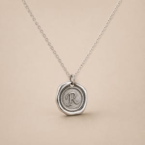 May include: A silver-toned necklace featuring a circular pendant with an irregular, wax-seal style edge. The center of the pendant displays a raised, embossed capital letter R. The pendant hangs from a delicate, thin silver-toned metal chain.