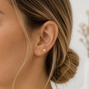 May include: A pair of small gold stud earrings worn in the earlobe. One earring is shaped like the capital letter S, and the other earring is shaped like a simple, solid heart. Both earrings have a polished, smooth finish and a minimalist design.