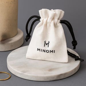以下が含まれることがあります： 黒い紐が付いた、オフホワイトの巾着袋。袋には黒字で「MINOMI」と書かれています。白い大理石のような表面に置かれています。左下には金色のリングが見えます。