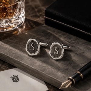 May include: A pair of silver-toned cufflinks featuring a circular wax seal design with a textured gray center and the letter S embossed in the middle. The cufflinks have a polished metallic border and a standard toggle closure mechanism on the back.
