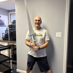 May include: A person holding a large, silver-coloured sword. The sword has a detailed hilt and a long, pointed blade. The person is wearing a grey t-shirt and dark shorts.