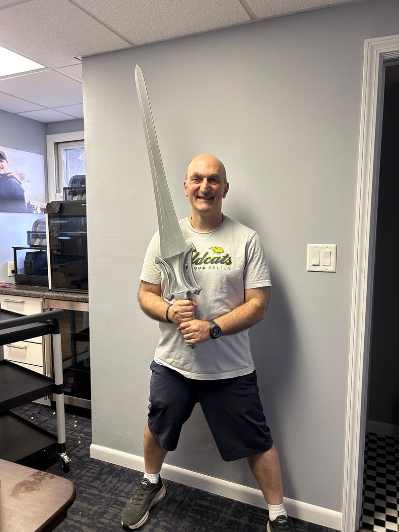 May include: A person holding a large, silver-coloured sword. The sword has a detailed hilt and a long, pointed blade. The person is wearing a grey t-shirt and dark shorts.