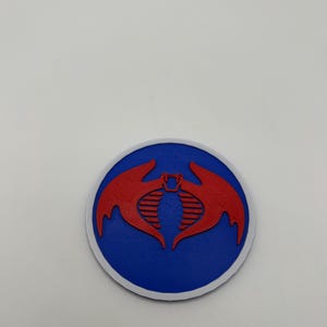 May include: A round emblem featuring a red cobra design on a blue background, with a white border. The cobra has a stylized open mouth and wings. The emblem is likely a logo or patch.