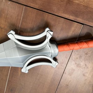 May include: A silver sword with a detailed hilt and an orange, wrapped handle. The blade has a geometric design. The sword is on a wooden surface.