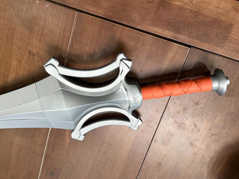 May include: A silver sword with a detailed hilt and an orange, wrapped handle. The blade has a geometric design. The sword is on a wooden surface.