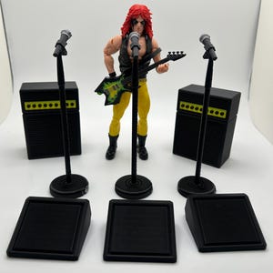 May include: A rock star action figure with red hair, a black vest, and yellow pants, holding a guitar. The figure is surrounded by black microphone stands, amplifiers, and stage platforms. The guitar has a green and yellow design.