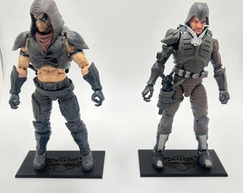 May include: Two action figures of soldiers in tactical gear. The figures are posed on black rectangular bases. One figure is primarily dark gray with a hooded cowl and a red neck covering. The other figure is in shades of gray and brown.