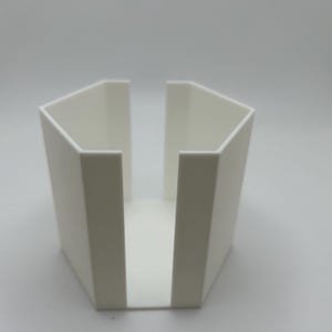 May include: A white, geometric, open-top container with a pentagonal shape. The sides are angled, and the front is open, creating a slot. The object is made of a solid, opaque material and sits on a white surface.