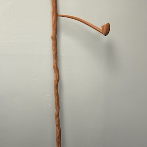 May include: A brown wooden staff with a carved top and a pipe-like attachment. The staff has a textured, naturalistic appearance, suggesting it is made to resemble a tree branch. The pipe is angled and has a small bowl.