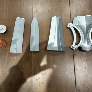 May include: A collection of 3D-printed prop weapon components in shades of grey and orange. The parts include a circular piece, a cylindrical handle, two long rectangular pieces, a blade-shaped piece, and a decorative shield-like element.