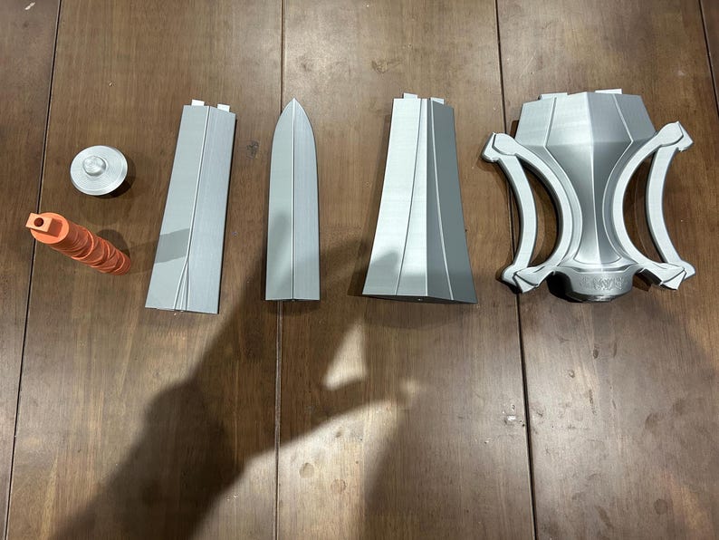 May include: A collection of 3D-printed prop weapon components in shades of grey and orange. The parts include a circular piece, a cylindrical handle, two long rectangular pieces, a blade-shaped piece, and a decorative shield-like element.