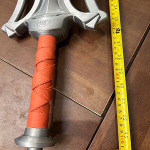 May include: A silver and orange sword prop, approximately 110 cm long. The sword features a detailed silver hilt and a wrapped orange handle. The blade has a unique design. The sword is lying on a wooden surface.