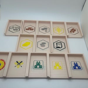 May include: A set of 14 wooden game tile holders with various symbols printed on them. The symbols include a sheep, bricks, a cloud, wheat, wood, a piece of paper, coins, a cat, a target, crossed swords, a castle in green, a castle in yellow, and a castle in blue.