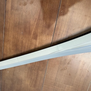May include: A curved, light grey object with a pointed end, possibly a component of a larger structure. The object has a smooth surface and is set against a wooden background. The design features a subtle, layered construction.