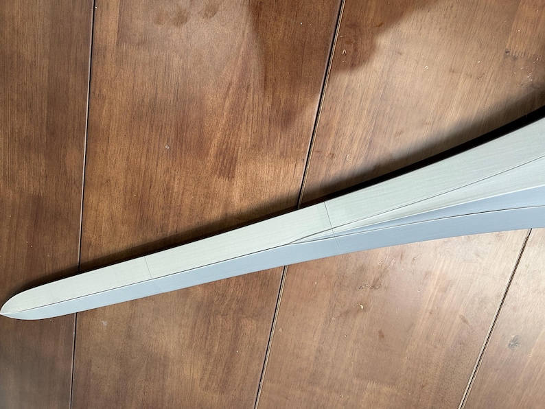 May include: A curved, light grey object with a pointed end, possibly a component of a larger structure. The object has a smooth surface and is set against a wooden background. The design features a subtle, layered construction.