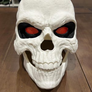 May include: A white skull mask with black eye sockets and red mesh eyes. The mask has detailed bone structure and a wide open mouth with visible teeth. The mask is likely for Halloween or costume use.