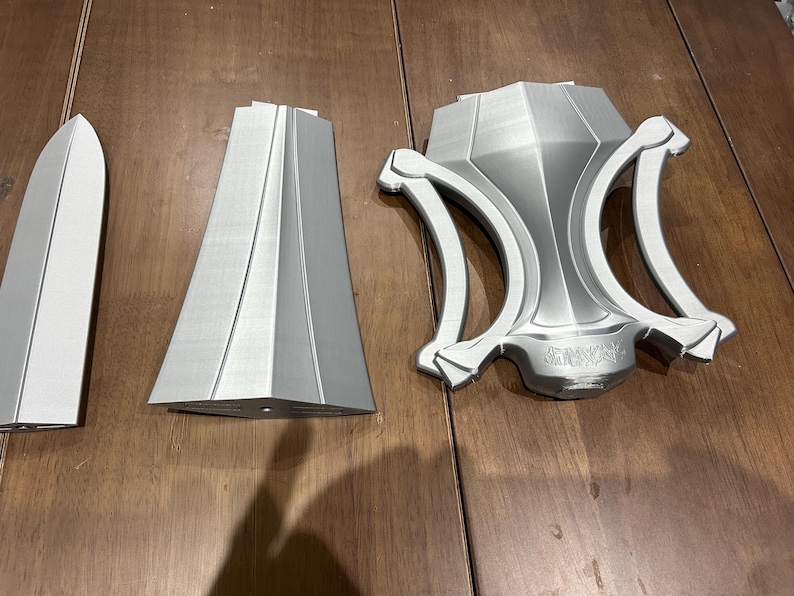 May include: A silver sword, in three parts, displayed on a wooden surface. The components include a blade, two side pieces, and a decorative hilt. The sword is unassembled.