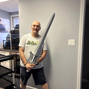 May include: A large, silver and grey sword with a detailed hilt is held by a person. The sword has a long, straight blade and ornate handle. The person is wearing a white t-shirt with the text "Wildcats NEUQUA" and black shorts.
