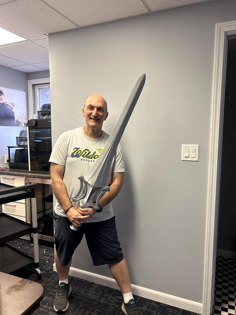 May include: A large, silver and grey sword with a detailed hilt is held by a person. The sword has a long, straight blade and ornate handle. The person is wearing a white t-shirt with the text "Wildcats NEUQUA" and black shorts.