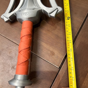 May include: A silver fantasy sword with an orange wrapped handle. The sword is approximately 112 cm long. The word "SAZAR" is engraved on the hilt. The sword has a decorative, ornate design.
