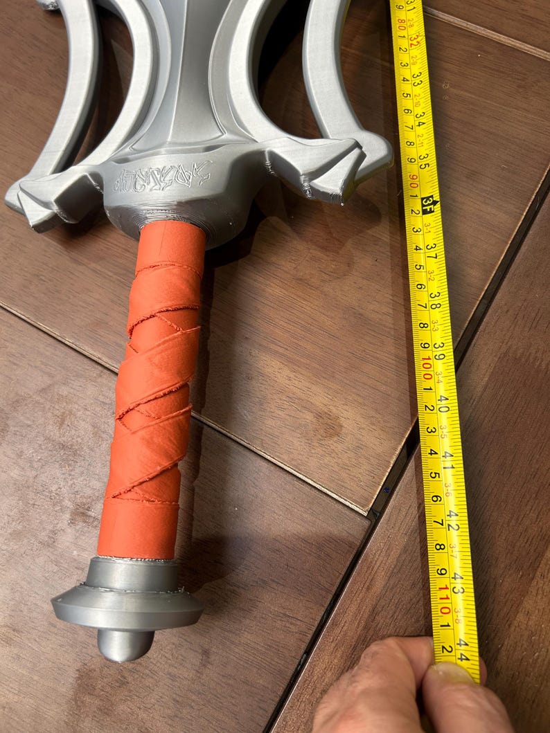 May include: A silver fantasy sword with an orange wrapped handle. The sword is approximately 112 cm long. The word "SAZAR" is engraved on the hilt. The sword has a decorative, ornate design.