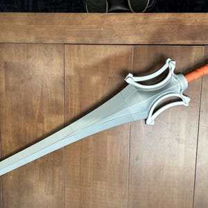 May include: A silver sword with a detailed hilt and an orange grip. The blade is long and pointed, with a series of geometric shapes. The sword is lying on a wooden surface.