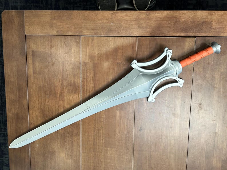 May include: A silver sword with a detailed hilt and an orange grip. The blade is long and pointed, with a series of geometric shapes. The sword is lying on a wooden surface.