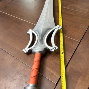 May include: A silver-coloured sword with a red wrapped handle. The sword is approximately 107 cm long. The sword has a detailed hilt and a long, pointed blade. The sword is lying on a brown wooden surface.