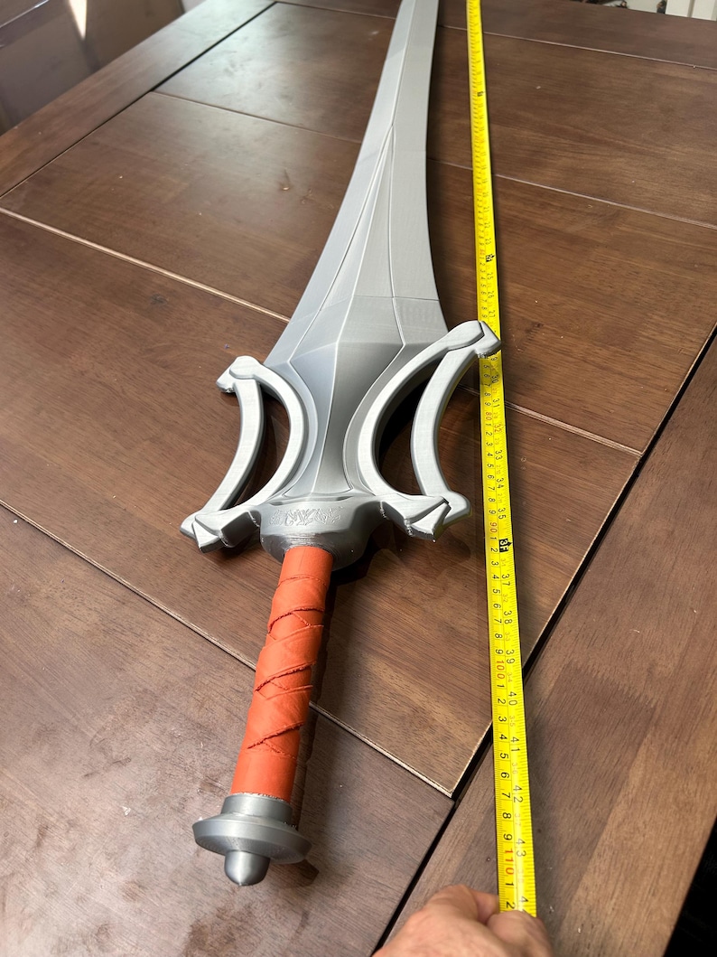 May include: A silver-coloured sword with a red wrapped handle. The sword is approximately 107 cm long. The sword has a detailed hilt and a long, pointed blade. The sword is lying on a brown wooden surface.