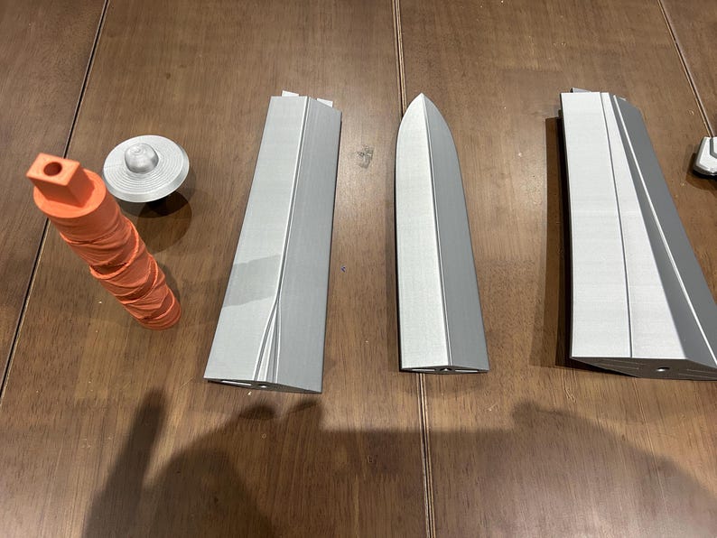 May include: A collection of 3D-printed parts. There's an orange, textured cylinder, a silver disc, and several grey, angular components. These could be elements of a larger model or project.