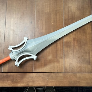 May include: A silver-colored sword with a long, pointed blade and a detailed hilt. The handle is wrapped in orange material. The sword is lying on a wooden surface.